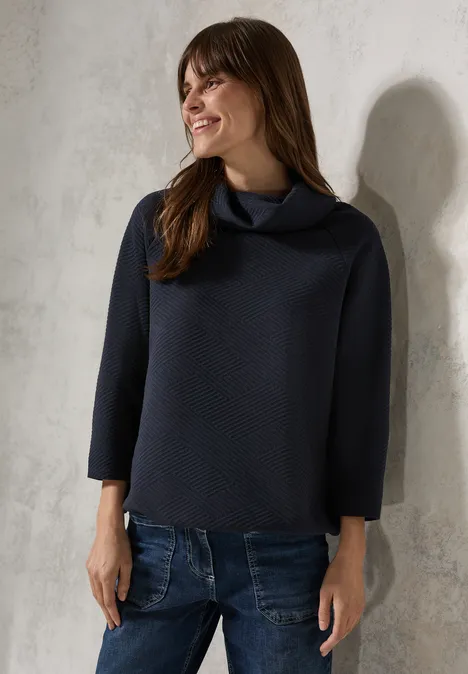 Frau mit dunklem Pullover und Jeans lächelt, steht vor heller Wand.